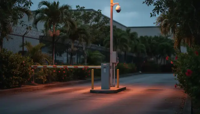 A barrier arm gate, card reader post and dome camera at the vehicle entrance of a gated commercial property at dusk